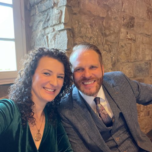 Ferienhaus Kellerwald Lächelndes Paar mit lockigem Haar und Anzug vor einer Steinmauer Ferienhaus Edersee, Gastgeber