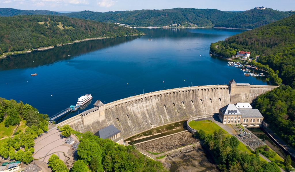 Ferienhaus Kellerwald Luftaufnahme eines Staudamms am See mit Boot und bewaldeten Hügeln im Hintergrund Ferienhaus Edersee, Partnerschaft mit edersee.com